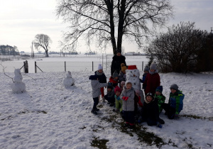 Na zdjęciu znajdują się przedszkolaki z pomocą nauczyciela oraz bałwan ulepiony przez dzieci, w tle widać zimowy krajobraz